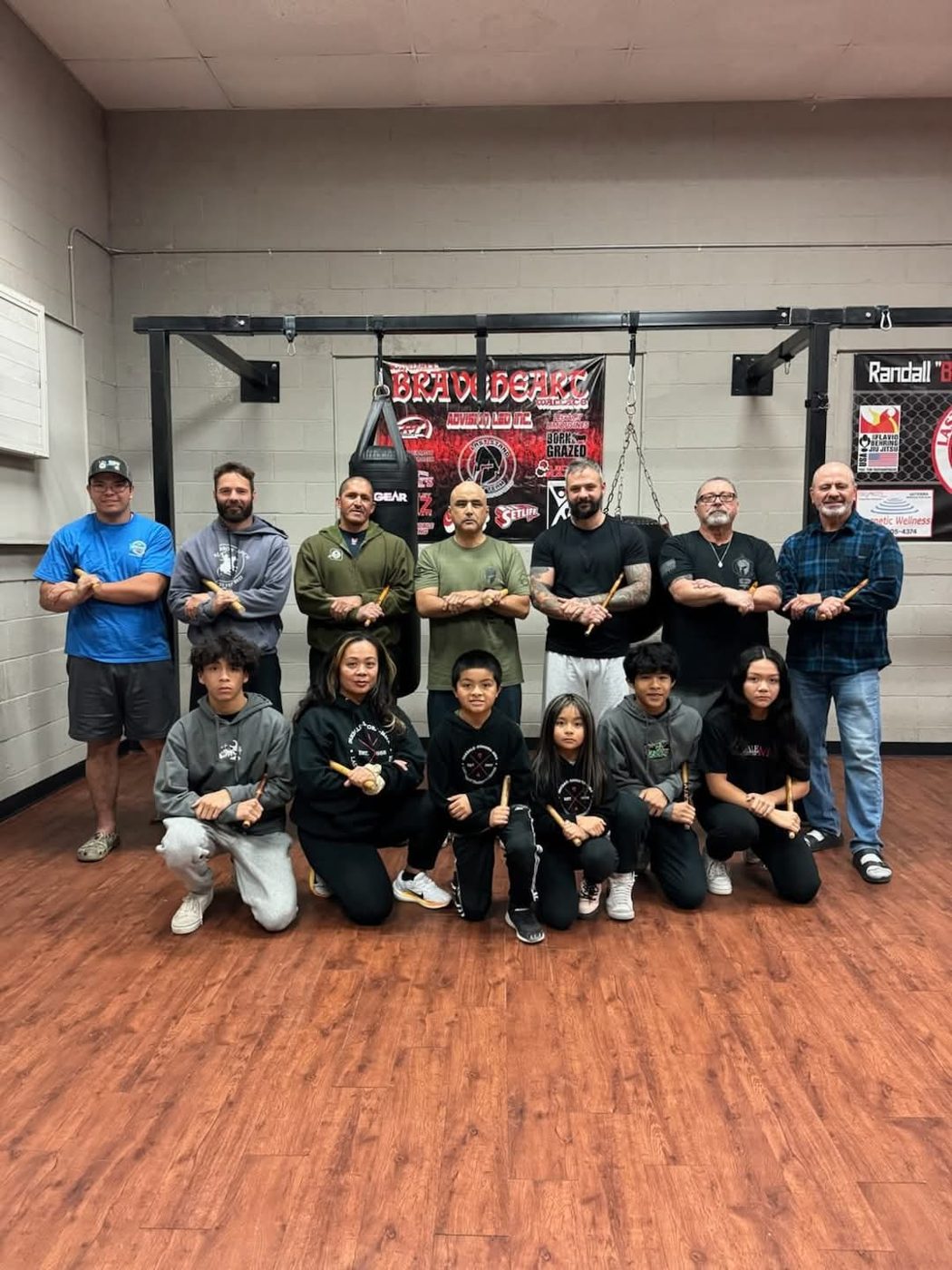 Escrima class of adults and teen students posing with training sticks in a martial arts gym
