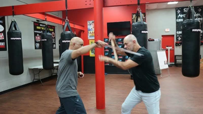 Master Edward Garcia in a gray shirt blocking a stick strike from a training partner during an Escrima drill